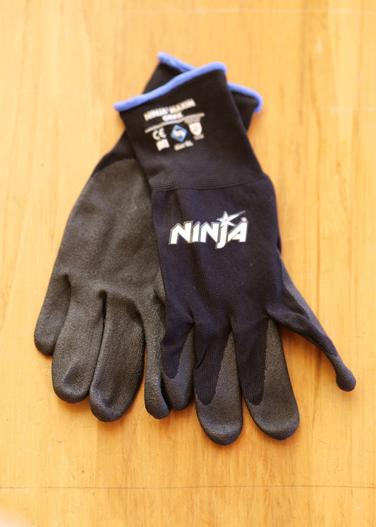 A pair of black and dark gray work gloves with blue trim and a white 'NINJA' logo, laid flat on a light wood surface.