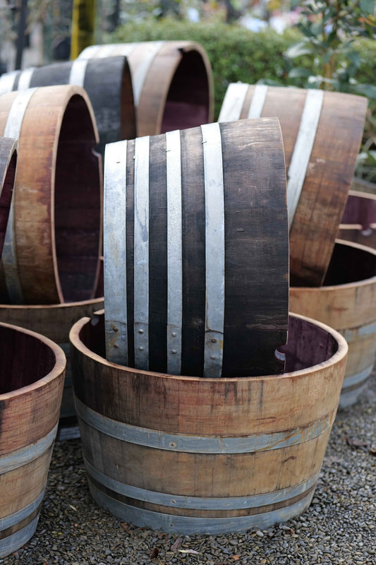 A close-up of a wooden wine barrel planter or bucket with dark stained wood and silver metal bands, with other similar barrels stacked in the background outdoors.